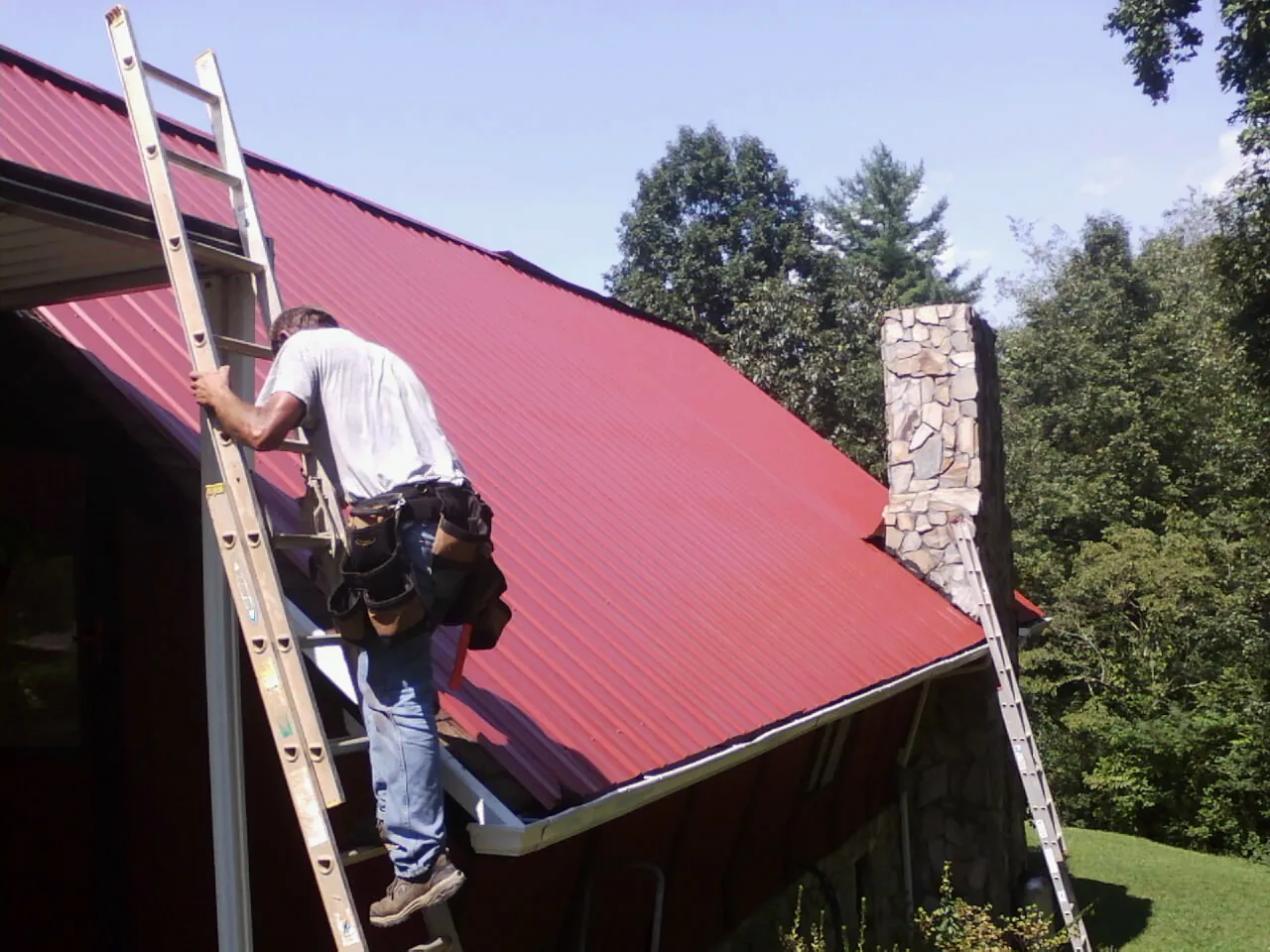 Roofer on a ladder beside a red metal roof and stone chimney — Soffit & Fascia Repair by Roofing Brothers in Bantam